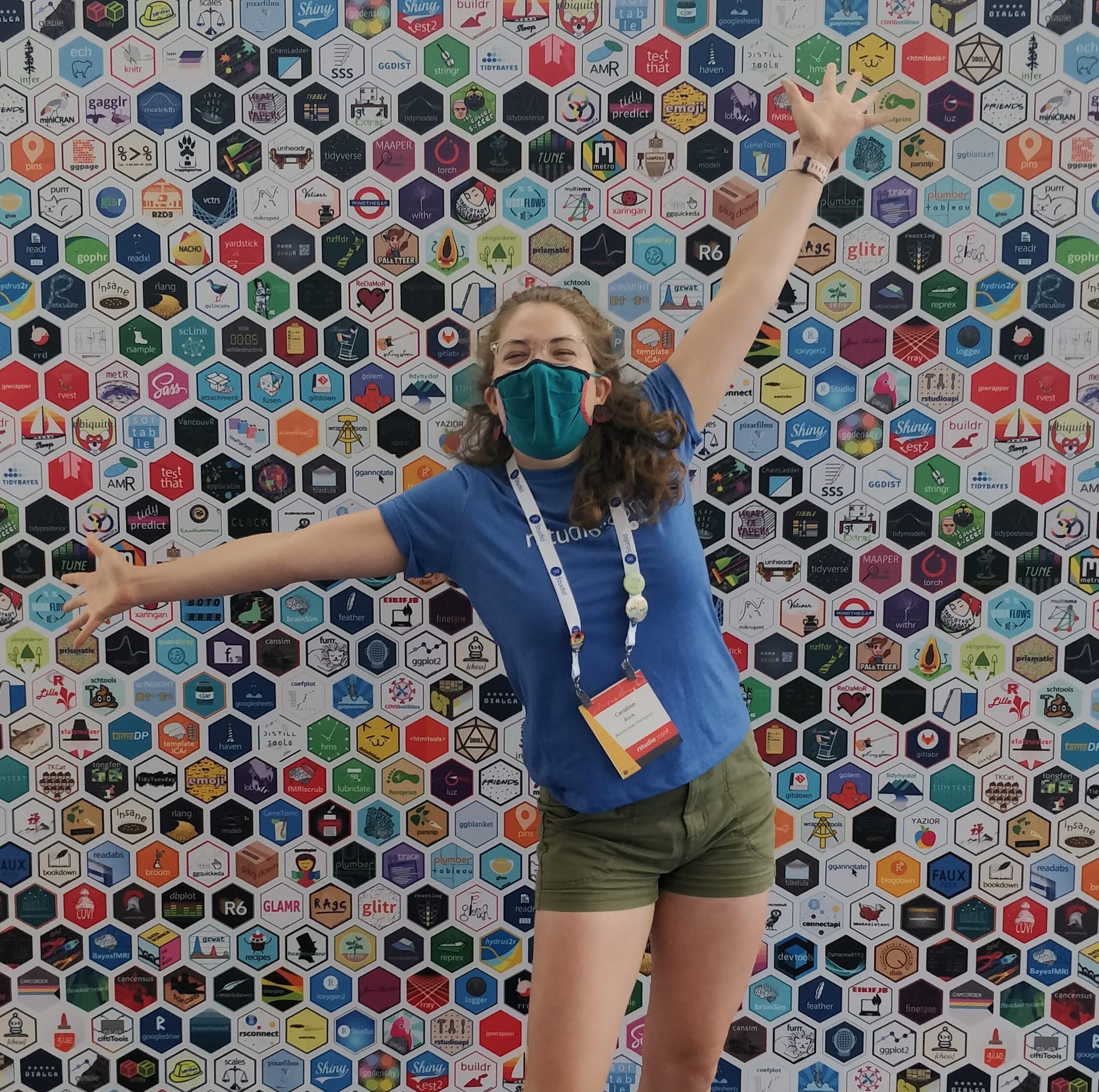 young woman standing in front of backdrop covered with R package hex logos. She looks very excited to be there.