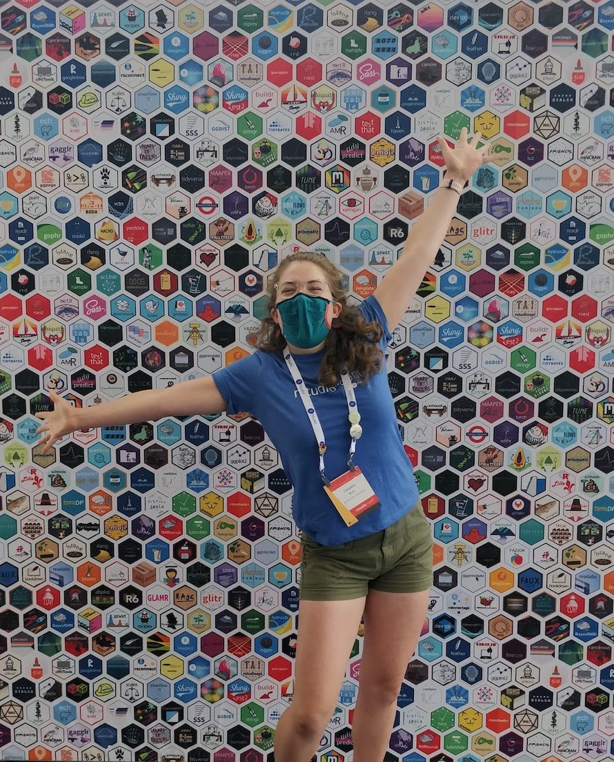 young woman standing in front of backdrop covered with R package hex logos. She looks very excited to be there.