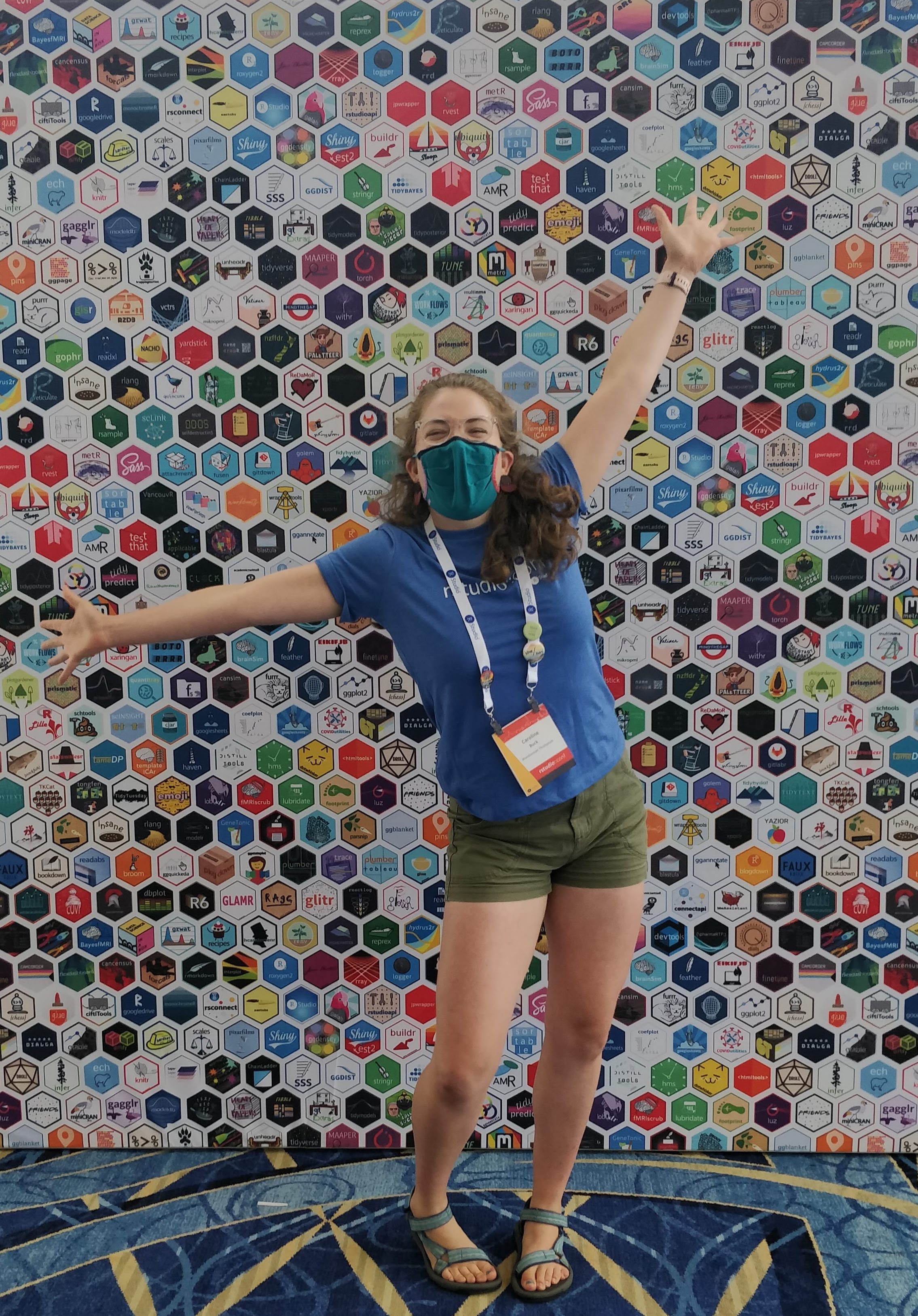 picture of a girl in blue Tshirt and green shorts standing excitedly in front of a background covered in R package stickers